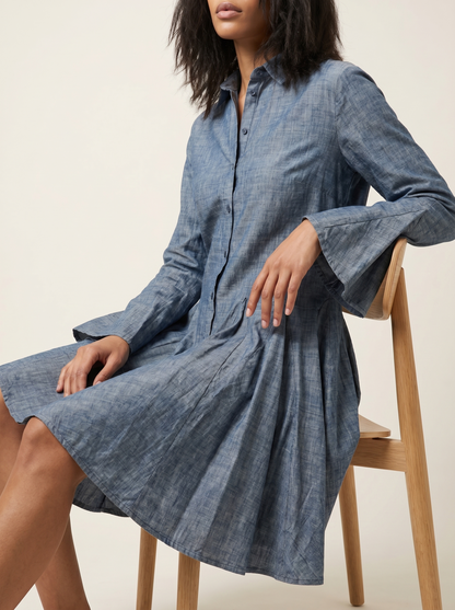 Woman wearing a blue denim dress sitting on a wooden stool against a beige background